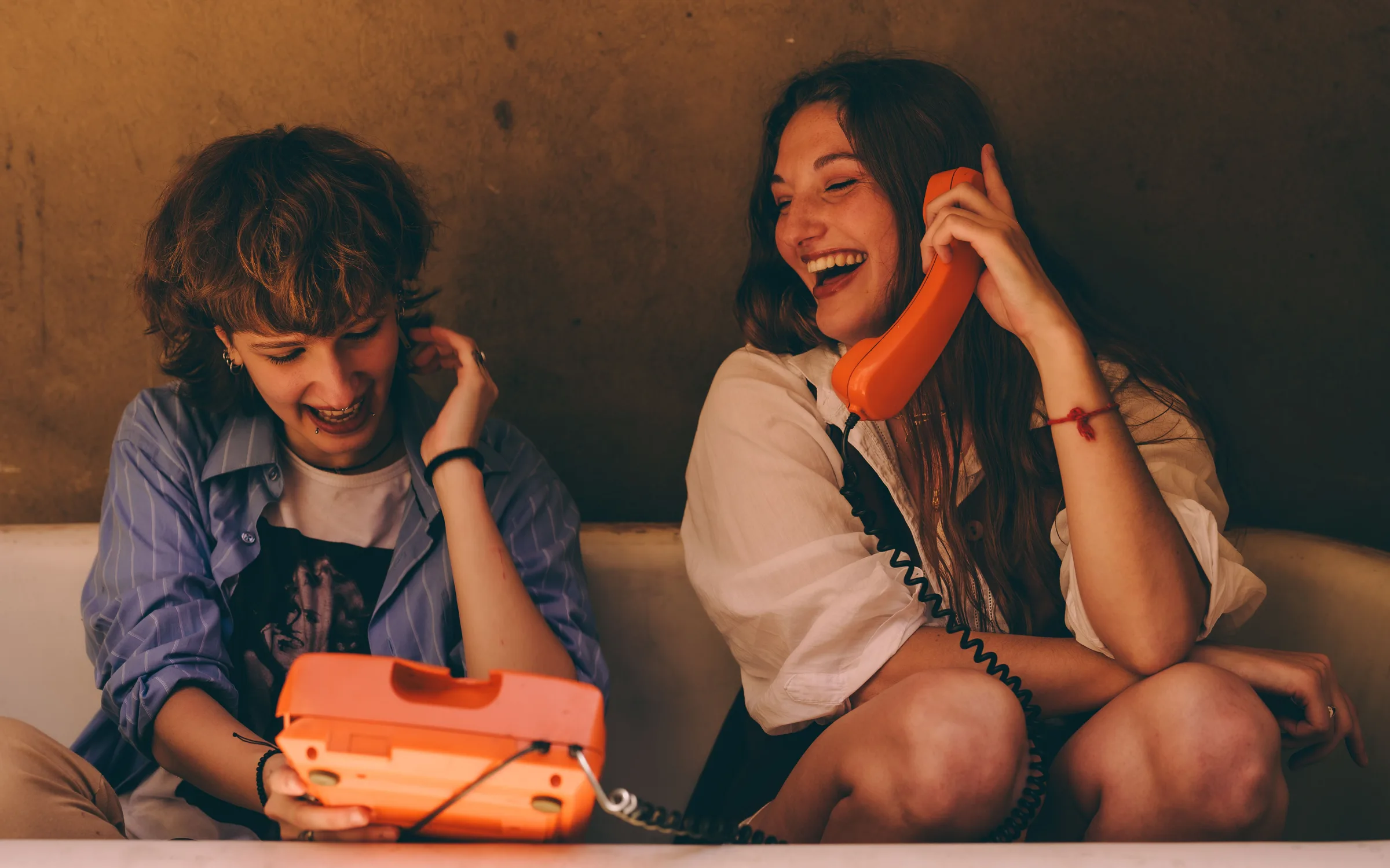 Deux amies rient ensemble, l’une tenant le combiné orange d’un vieux téléphone, l’autre le socle posé sur ses genoux.